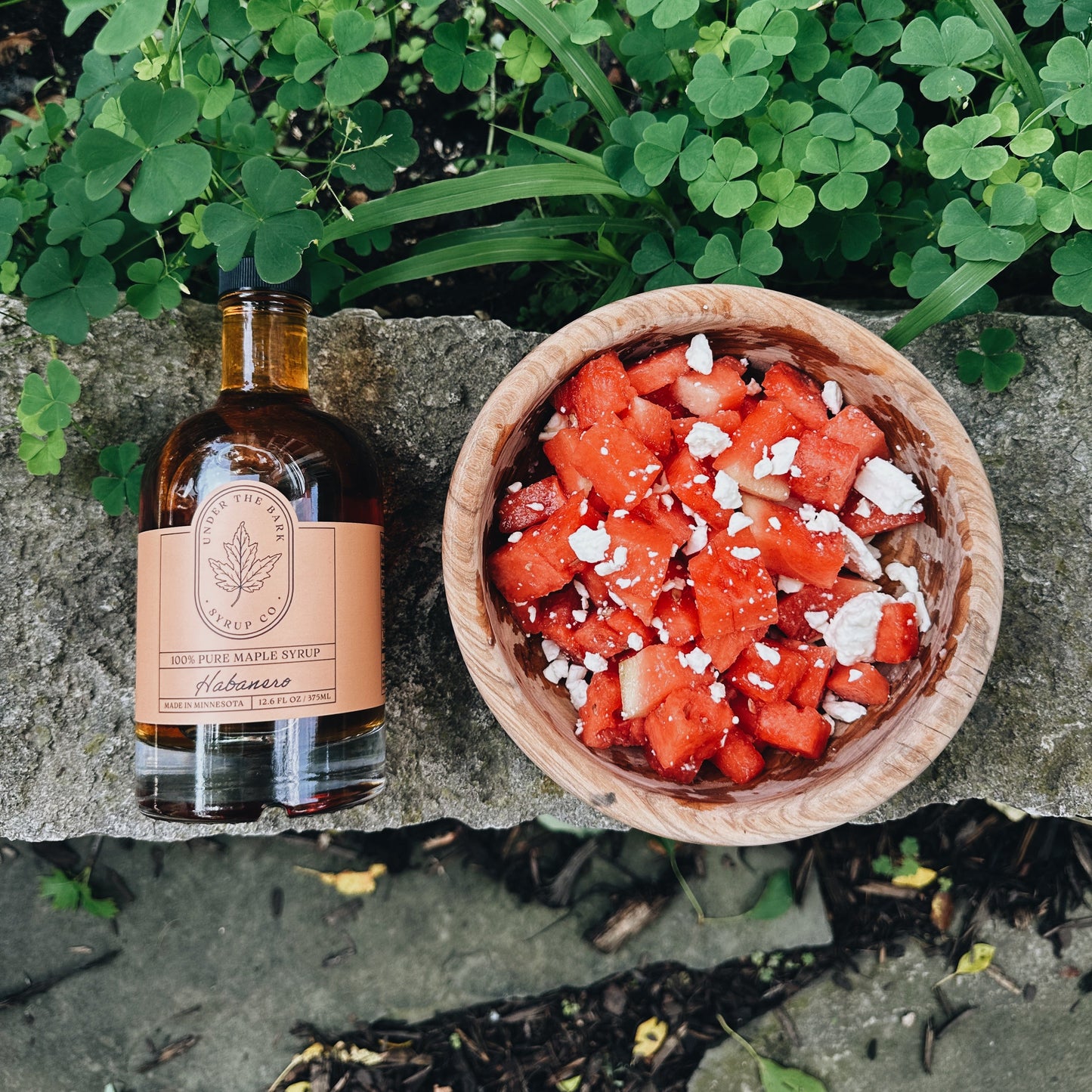 
                  
                    Bottle of habanero maple syrup and a bowl of watermelon salad on a stone surface with greenery in the background
                  
                