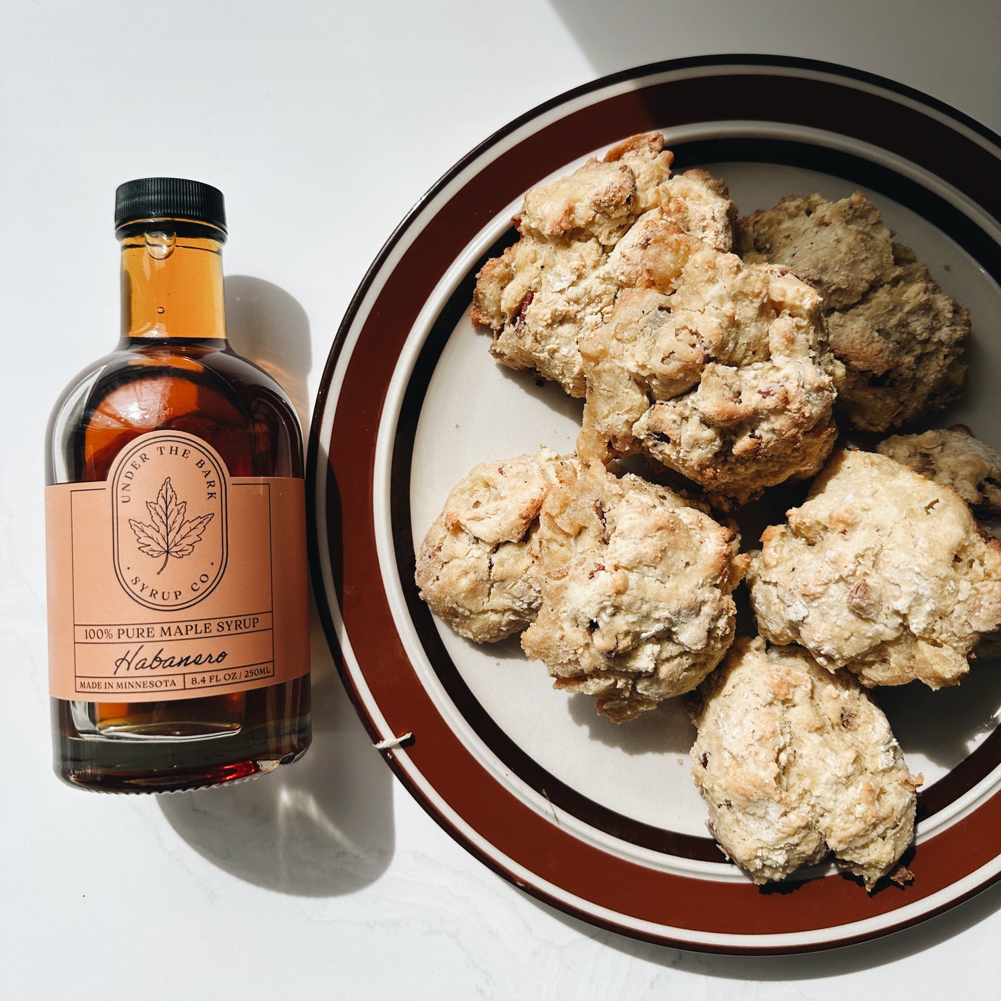 
                  
                    Bottle of habanero maple syrup next to a plate of biscuits on a white table.
                  
                