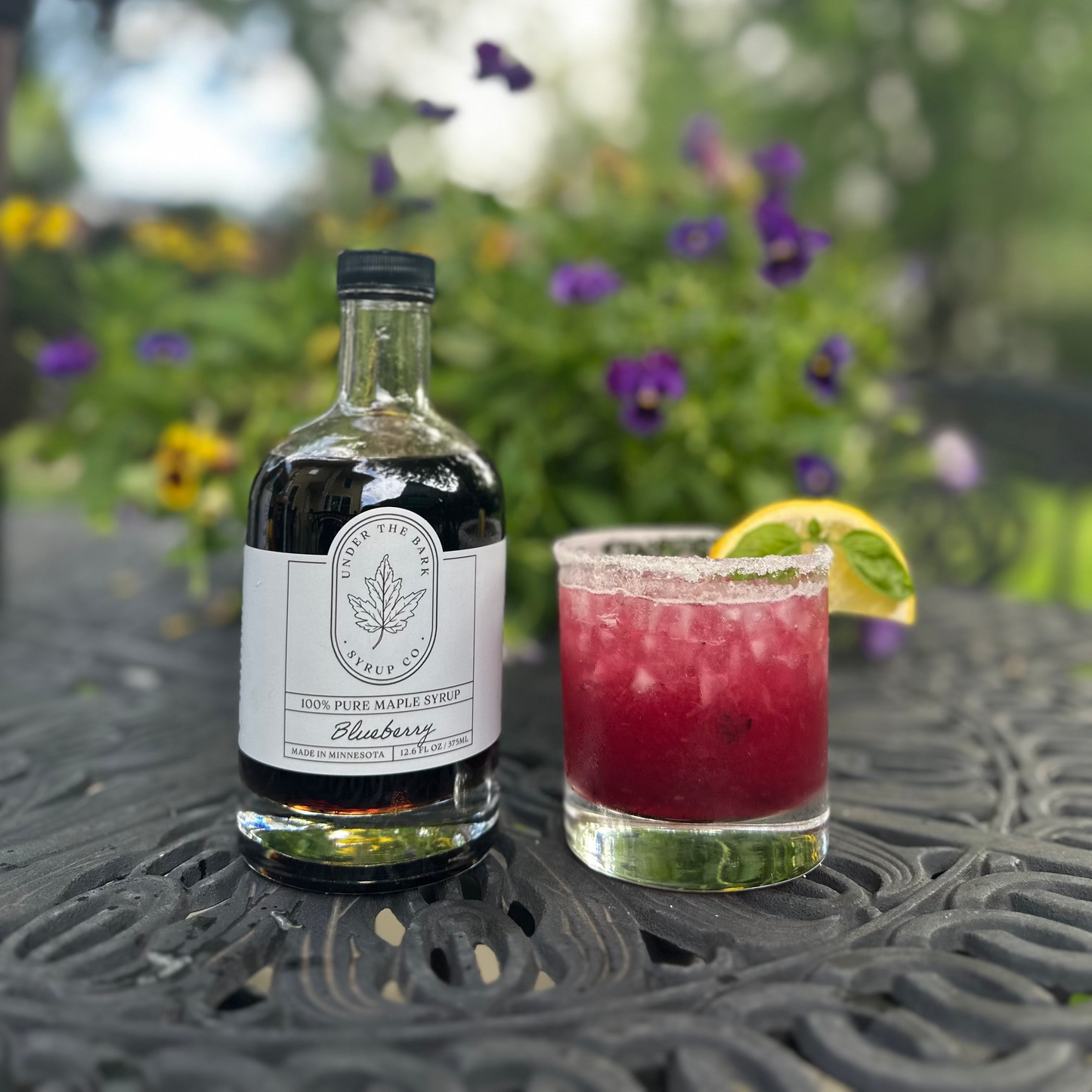 
                  
                    Bottle of blueberry liqueur and a cocktail on a table with a blurred garden background
                  
                