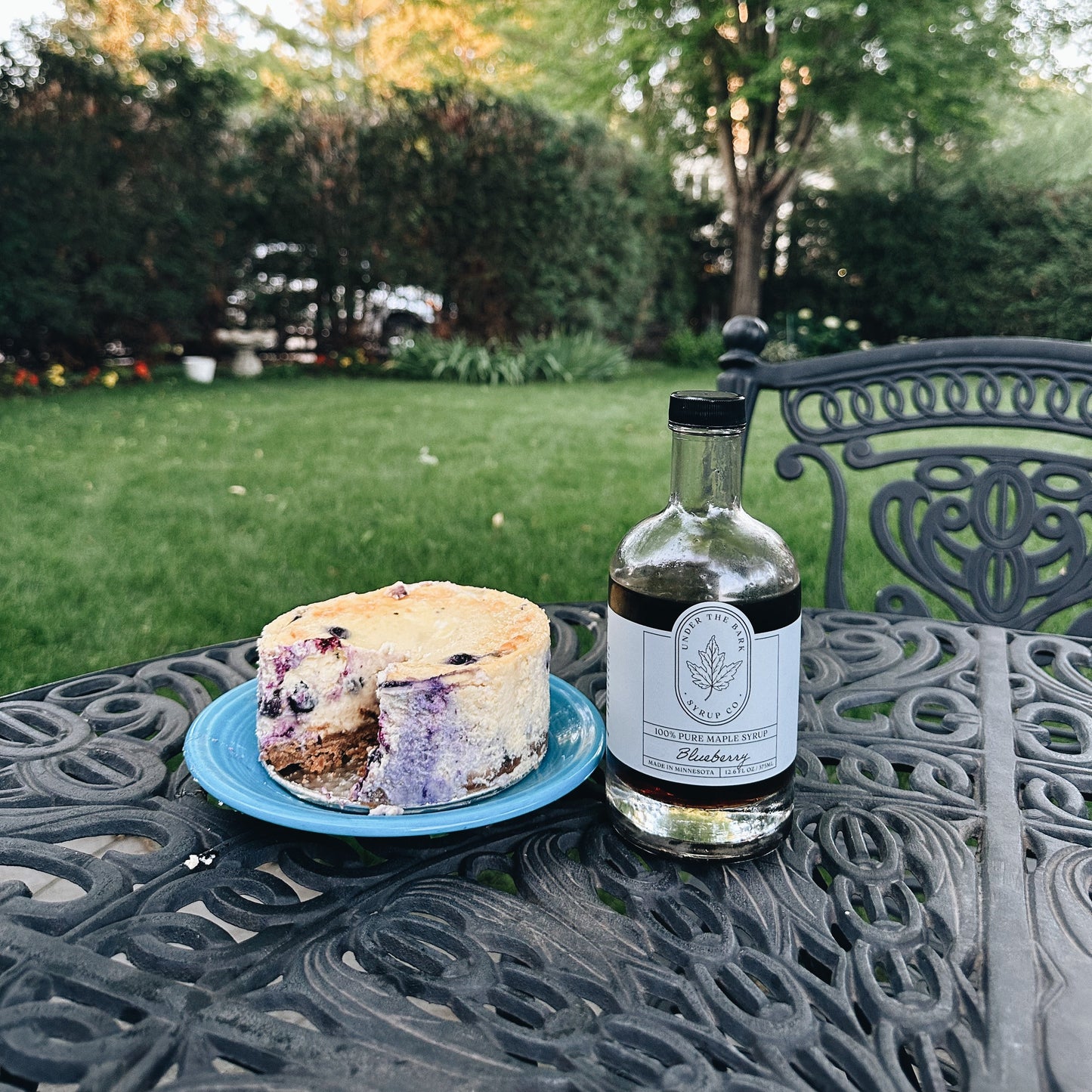 
                  
                    Bottle of blueberry maple syrup and a cake on a patio table with a garden background
                  
                